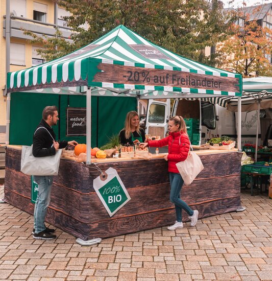 Der Marktstand ist vollflächig bedruckt und kommt auf dem Wochenmarkt und Bauernmarkt zum Einsatz. 2 Kunden sehen sich die Produkte an.