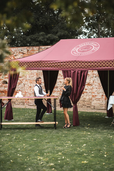 Im großen Garten steht das bordeaux-farbene Cateringzelt. Die Frau und der Mann haben ein Sektglas in der Hand und stehen an einem Stehtisch.