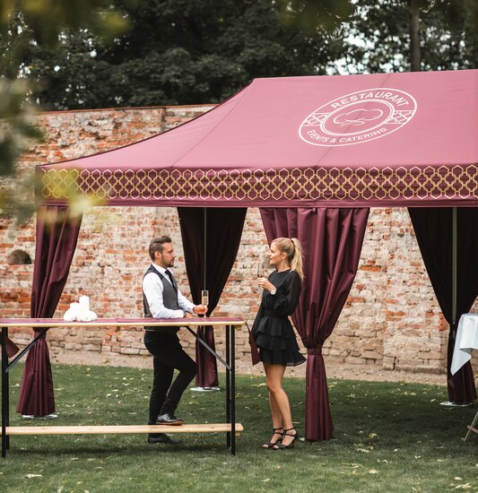 The bordeaux-coloured catering tent stands in the large garden. The woman and the man have a champagne glass in their hands and are standing at a high table.