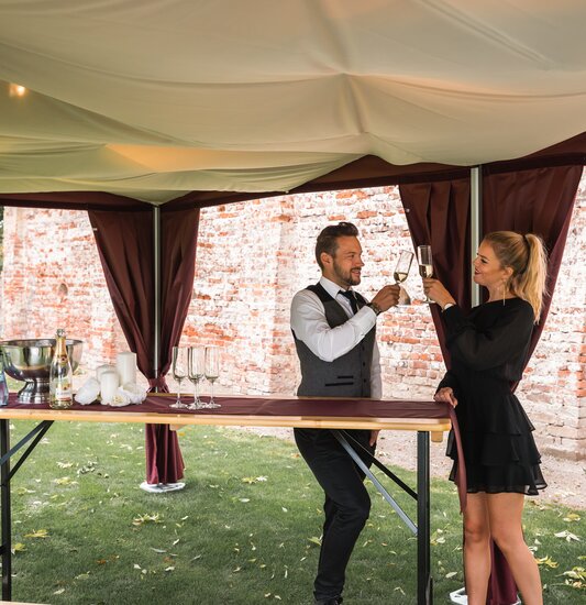 Unter dem eleganten Faltpavillon mit Himmel stehen 2 Personen mit einem Glas Prosecco in der Hand. 