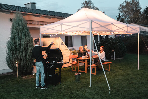 Freunde feiern unter dem Gartenzelt eine Grill- und Gartenparty. Das Gartenzelt ist feierlich dekoriert mit Lichterketten.