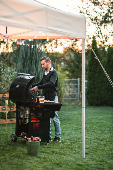 The man is standing with his barbecue under an ecru-coloured folding gazebo in the garden. The folding gazebo is decorated with fairy lights and secured with tension belts.