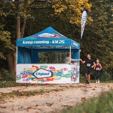 Blauer 3x3m Eventzelt mit individuellem Citylauf-Druck, mit individueller Fahne, geschlossener Seitenwand und halbhoher Seitenwand mit Theke als Verpflegungsstand bei einem Stadtlauf